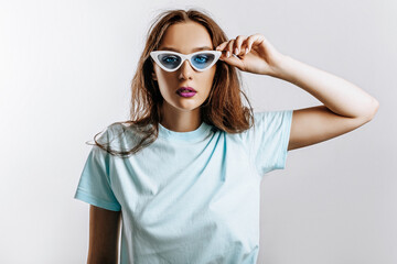 Beautiful young brunette girl with green eyes with fashion makeup with purple lips on an isolated gray background. The woman in blue sunglasses seriously looking at the camera. Beauty concept.