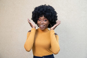 Portrait of delighted African American cheerful woman with curly hair and happy face, raises palms and looking at camera, reacts on awesome gift, posing on light background. People, ethnicity concept.