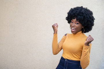 Joyful African American woman with curly hair keeps fists clenched, smiles broadly, celebrates success, wearing casual wear, looking at camera, posing on light background. Empty copy space.