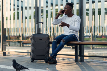 Handsome african-american male traveler with a suitcase calls a taxi on mobile phone sitting at bus stop.