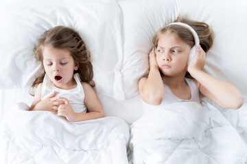 Little girls sleep in bed. One is snoring hard,sister is plugging ears with fur headphones.Early...