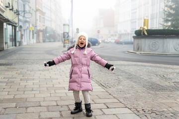 Fototapeta premium Cute liitle sad lost caucasian girl in warm long pink jacket and hat shouting loud on empty street of old european city Wels. Screaming angry alone scared child in town in Morning fog on backround