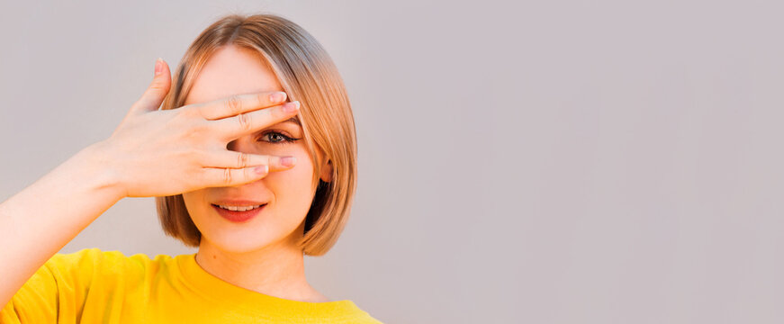 Glad smiling blonde female model hiding her face behind hand, has broad smile, happy to recieve compliments from handsome man, isolated against gray background. Positive young woman