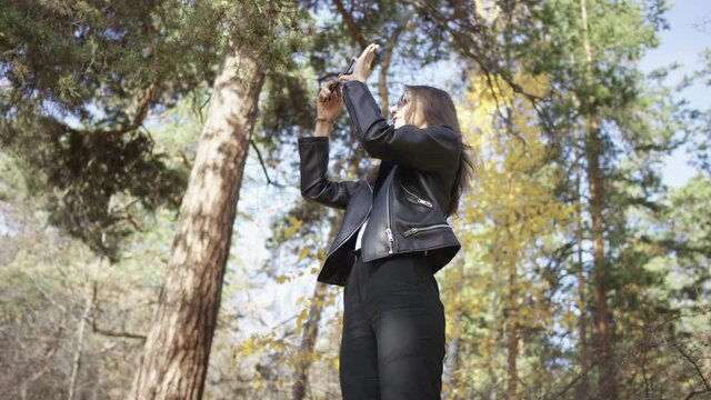 Good-looking female photographer in sunglasses takes pictures of beautiful autumn trees at sunny day with smartphone. Low angle 4K 360 degree tracking arc shot.