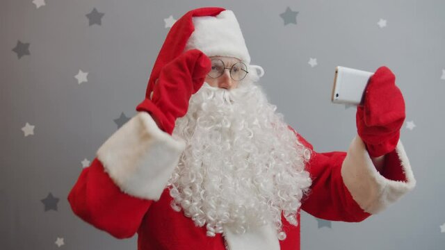 Portrait of Santa Claus taking selfie with smartphone camera on grey glittery background touching screen and looking at camera. Moern gadgets and holidays concept.