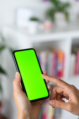 Mockup image of a woman's hand holding and using a mobile phone with blank green screen in living room