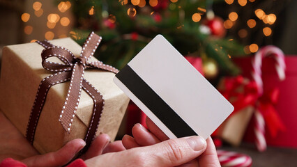 Young woman holding a credit card and a gift box against the background of Christmas decor and...