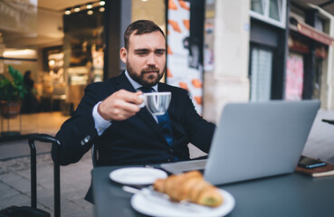 Fototapeta na wymiar Caucasian male entrepreneur with coffee cup watching webinar seminar during breakfast time in street cafe, formally dressed businessman reading financial news about trade exchange using laptop app