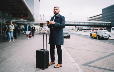 Male employee in formal clothing standing near airport pondering on departure during work travel, Caucasian entrepreneur with suitcase luggage holding cellphone technology and looking for terminal