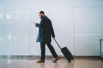 Elegant businessman checking e-mail on mobile phone while walking with suitcase in airport, formally dressed male employer using cell technology for booking return tickets from work travel
