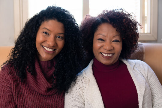 Portrait Of Brazilian Adult Daughter And Mother Sitting In Couch Looking At Camera Inside The House In Living Room. Family, Affectionate, Reunion, Together Concept..