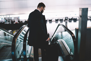 Back view of formally dressed male pilot going to electric stairs for getting to departure gate, businessman with luggage suitcase standing on escalator have work trip from airport terminal