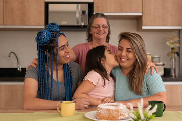 Joyful brazilian young child kissing one mother in the cheek in kitchen home, indoors. Lgbtq family, bonding, love, generation concept..