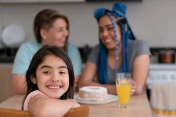 Portrait of brazilian young child with mothers smiling at kitchen table, inside. Adoption, same sex family, parenting concept..