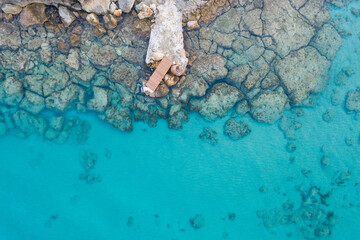 An aerial view of the beautiful Mediterranean Sea, with a wooden pier and a rocky shore, where you can see the textured underwater corals and the clean turquoise water of Protaras, Cyprus	