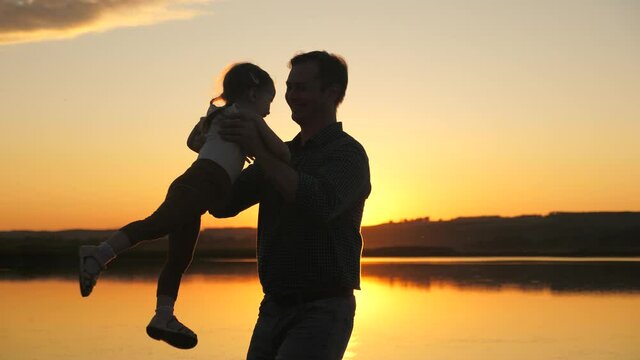 silhouette of a father dancing with toddler in sun. Daddy is spinning with his daughter in air on beach, having fun at sunset. father plays with child by water. happy family and childhood concept.