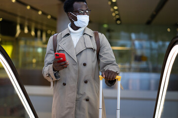 Afro-American traveler man with yellow suitcase stands on escalator in airport terminal, wear face medical mask to protect yourself from contact with flu virus, pandemic covid-19. New normal concept