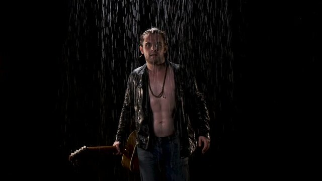 Cool muscular rocker in black leather jacket on naked body walks forward with guitar in his hand in the rain on black background. Concept for the final touch at the concert.