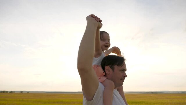 Dad carries on shoulders of his beloved child, in rays of sun. Father walks with his daughter on his shoulders in rays of sunset. child with parents walks at sunset. happy family resting in park.
