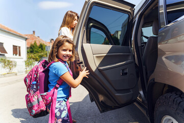 Little girl in sunny day or morning going to school - small pupil first grader with backpack on shoulders and her mother entering the car - Real people education concept