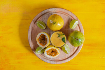 Top view of some passion fruit over wood plate and glass of passion fruit smoothie by fresh ingredients with lime and mint on yellow wooden background