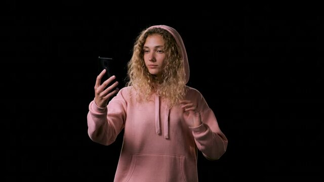 Beautiful nice woman in pink hoodie makes a selfie on a black background. A lady with long blond hair picks up the phone and looks into it, talks to a friend via video link.