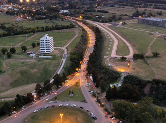 Vista aérea durante el atardecer de una ruta iluminada, con un edificio a la izquierda y una cruz con Cristo a la derecha.