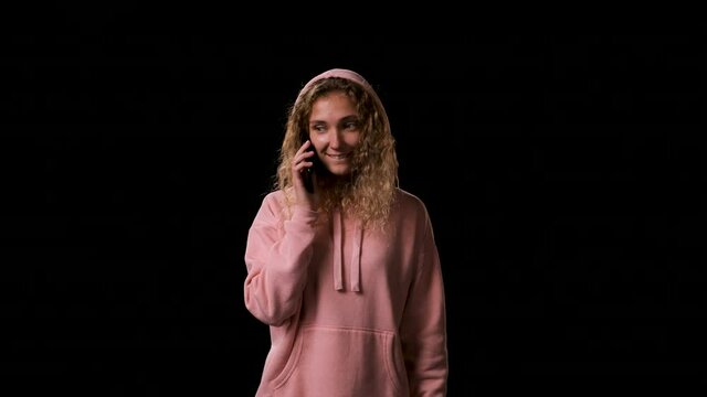 Beautiful lovely woman in pink sweatshirt with hood gets call on phone on black background. lady with long blond hair picks up the phone and hears very good news and is happy about it.