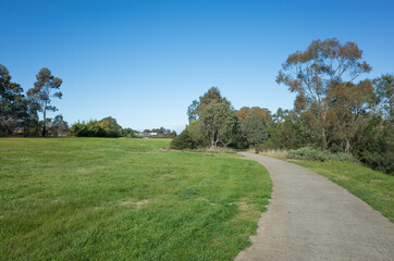 Fototapeta na wymiar A concrete footpath/walkway in a large park with vast lawn areas, Australian native Eucalyptus trees and some houses in the distance. Werribee River Trail, Melbourne VIC Australia.
