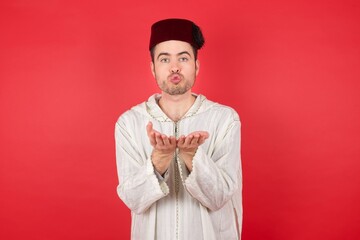 young caucasian muslim man wearing djellaba and traditional hat over red background sending blow...