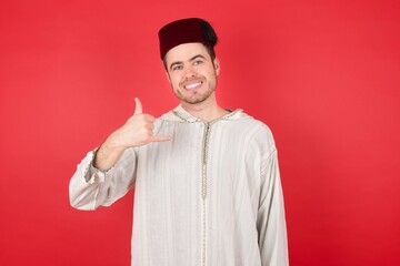 young caucasian muslim man wearing djellaba and traditional hat over red wall making call gesture with hands. Lovely 