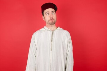 young caucasian muslim man wearing djellaba and traditional hat over red wall making fish face with lips, crazy and comical gesture. Funny expression.