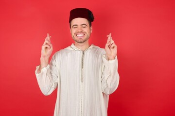 young caucasian muslim man wearing djellaba and traditional hat over red wall gesturing finger crossed smiling with hope and eyes closed. Luck and superstitious concept.