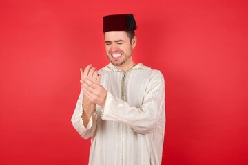 young caucasian muslim man wearing djellaba and traditional hat over red wall Suffering pain on hands and fingers, arthritis inflammation