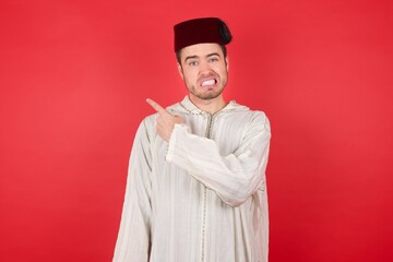 Portrait of displeased upset young caucasian muslim man wearing djellaba and traditional hat over red background Pointing aside worried and nervous with forefinger, concern and surprise concept.