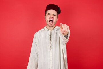Portrait of displeased upset young caucasian muslim man wearing djellaba and traditional hat over red wall pointing displeased and frustrated to the camera, angry and furious ready to fight with you.