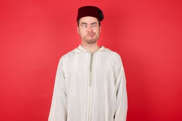 Portrait of displeased upset young caucasian muslim man wearing djellaba and traditional hat making grimace and crazy face, screaming out of control, funny lunatic expressing freedom and wild.