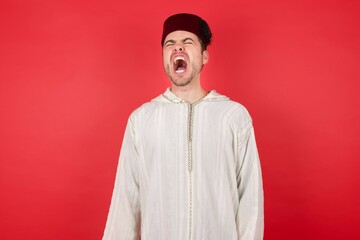 Obraz premium Portrait of displeased upset young caucasian muslim man wearing djellaba and traditional hat over red angry and mad screaming frustrated and furious, shouting with anger. Rage and aggressive concept.