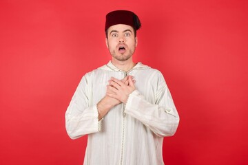 Scared young caucasian muslim man wearing djellaba and traditional hat over red background looks with frightened expression, keeps hands on chest, being puzzled to notice something strange.