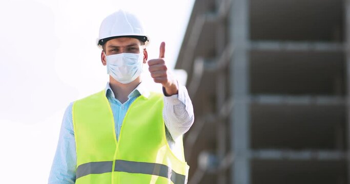 Professional builder in protective mask and helmet gesturing thumb up, standing at construction site