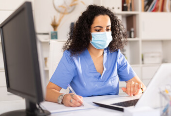 Female therapist in protective mask working at table with laptop in her office