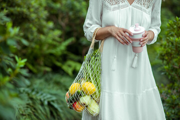 Woman in white dress with bags of vegetables and no-spill cup in hands