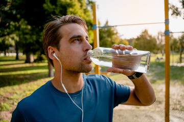 Thirsty young male athlete drinking water looking upwards while listening to music in ear phones...