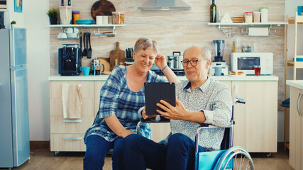 Senior couple laughing and waving during a video call with grandchildrens using tablet computer in kitchen. Paralysied handicapped old elderly man using modern communication techonolgy.