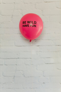 Words ""be wild have fun"" in black on pink balloon flying against white brick wall