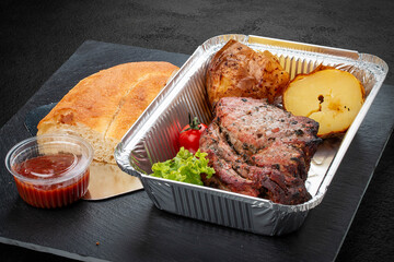 Grilled pork steak with baked potatoes, matnakash and red sauce. Food delivery. Served on aluminium lunch box.