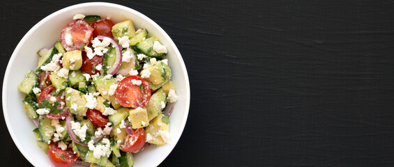 Delicious Avocado Tomato and Cucumber Salad in a white bowl on a black background, top view. Flat lay, overhead. Copy space.