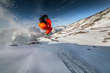 Wide angle male skier in orange suit makes a jump from a snowy ledge in the mountains. Snow powder trailing behind the athlete