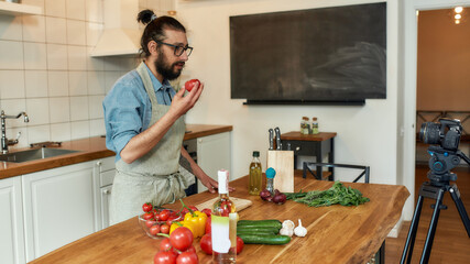 Young man Italian cook in apron looking at camera, holding tomato filming himself for culinary blog while preparing healthy meal with vegetables in the kitchen