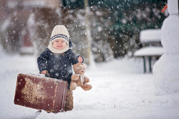 Baby playing with teddy in the snow, winter time. Little toddler boy in blue coat, holding suitcase...